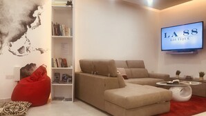 Una sala de estar moderna con un sofá color beige, un sillón de peluche rojo, una estantería y un televisor que muestra 'LA 88 BOUTIQUE'.
