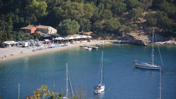A coastal area with a beach, boats, and a building with multiple umbrellas. - Villa Fedra (Corfu)