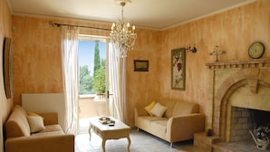A living room with two sofas, a coffee table, a chandelier, a fireplace, and a large window with curtains. - Villa Fedra (Corfu)