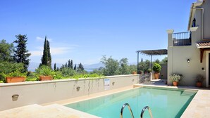 A swimming pool with a clear view of the mountains in the background, surrounded by potted plants and a building with a balcony. - Villa Fedra (Corfu)