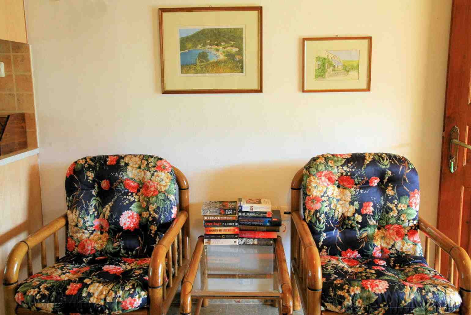 Deux fauteuils en bois recouverts d’un tissu à motifs floraux, une petite table avec des livres et deux tableaux encadrés accrochés au mur.