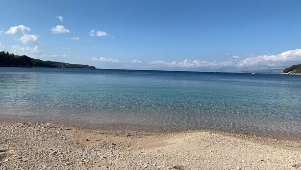 A clear view of a beach with pebbles and calm water under a blue sky. - Cochelli Apartments (Corfu)