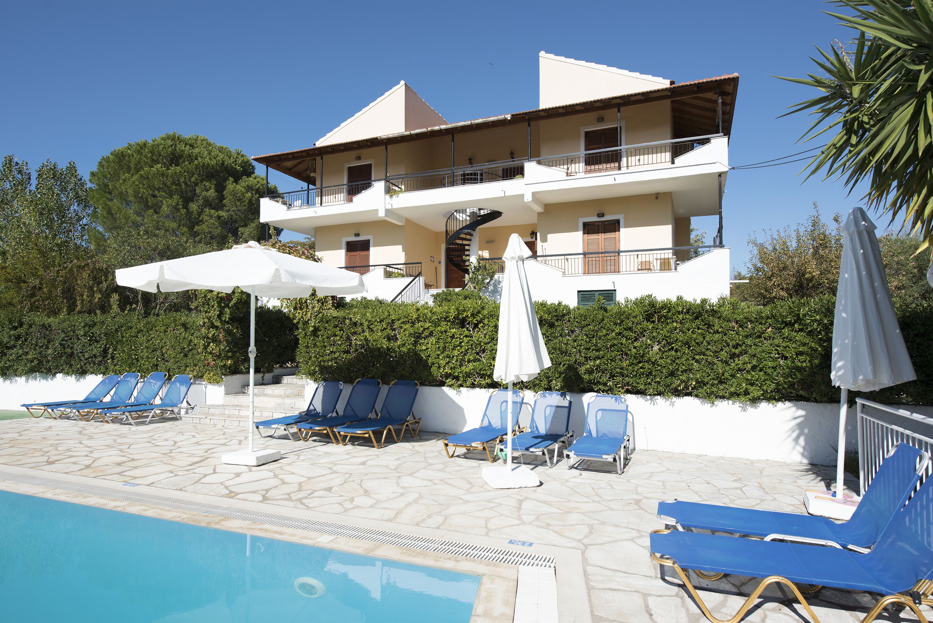 A pool area with lounge chairs and umbrellas, a multi-story building with balconies, and a clear blue sky.