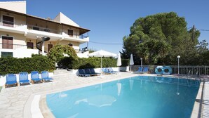 A swimming pool with lounge chairs and umbrellas, surrounded by a building with balconies and a tree. - Cochelli Apartments (Corfu)