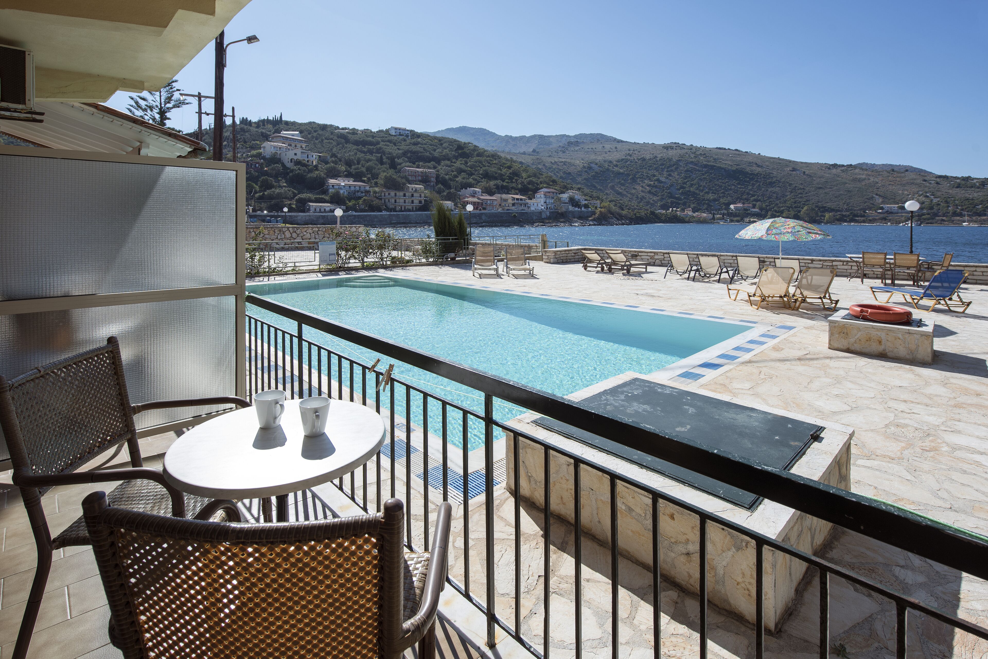 A poolside area with a view of the sea and hills, wicker chairs, a table with cups, and a lifebuoy.