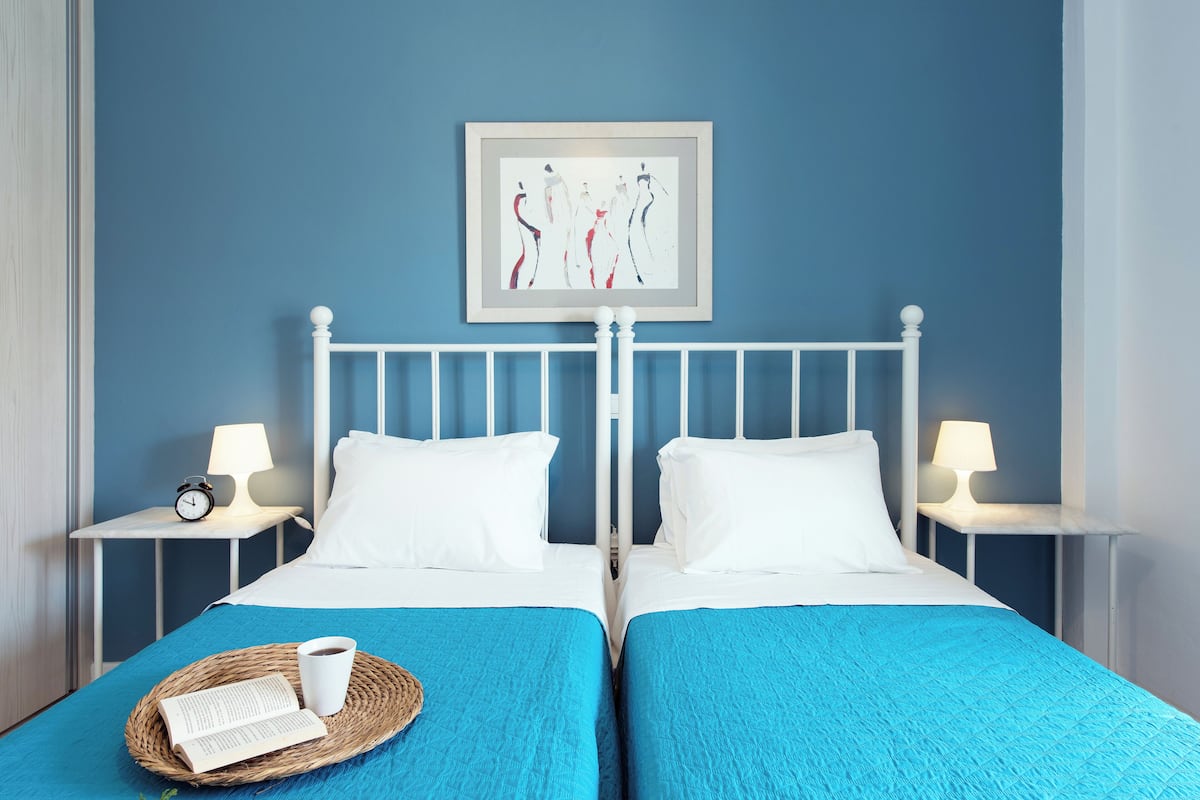 Ein Doppelbett mit weißen Kopfteil und Fußteil, zwei Nachttische mit Lampen, ein gerahmtes Kunstwerk über dem Bett und ein Tablett mit Buch und Tasse auf dem Bett.