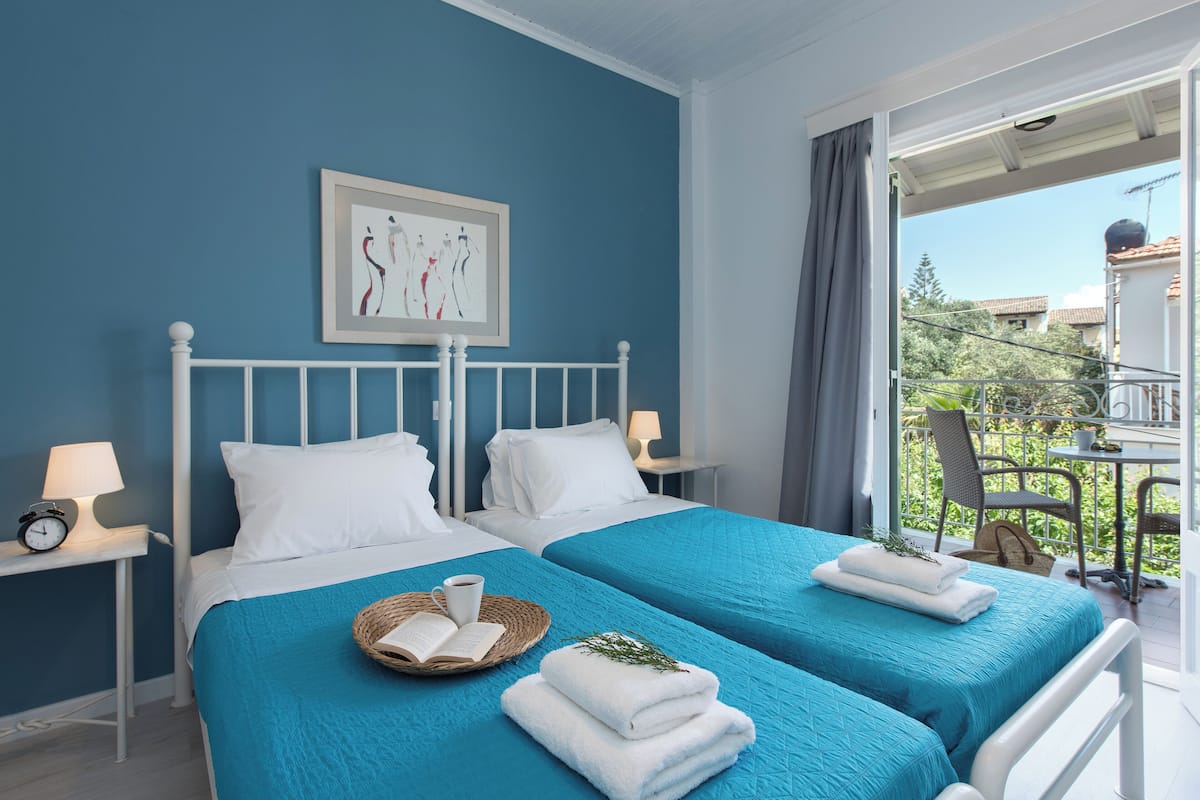 Ein Hotelzimmer mit zwei Betten, einem gerahmten Kunstwerk an der Wand, einem Balkon mit Blick auf Grünflächen und Gebäude sowie einem kleinen Tisch mit Lampe und Korb.