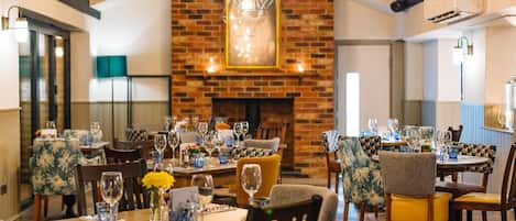 Un restaurant doté de nombreuses tables dressées pour le service, d’une cheminée en briques et d’un éclairage moderne.