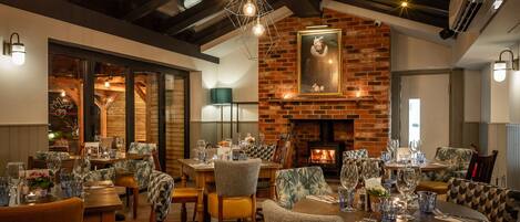 A dining area with brick walls, a fireplace, and modern lighting fixtures. Tables are set with wine glasses and cutlery.