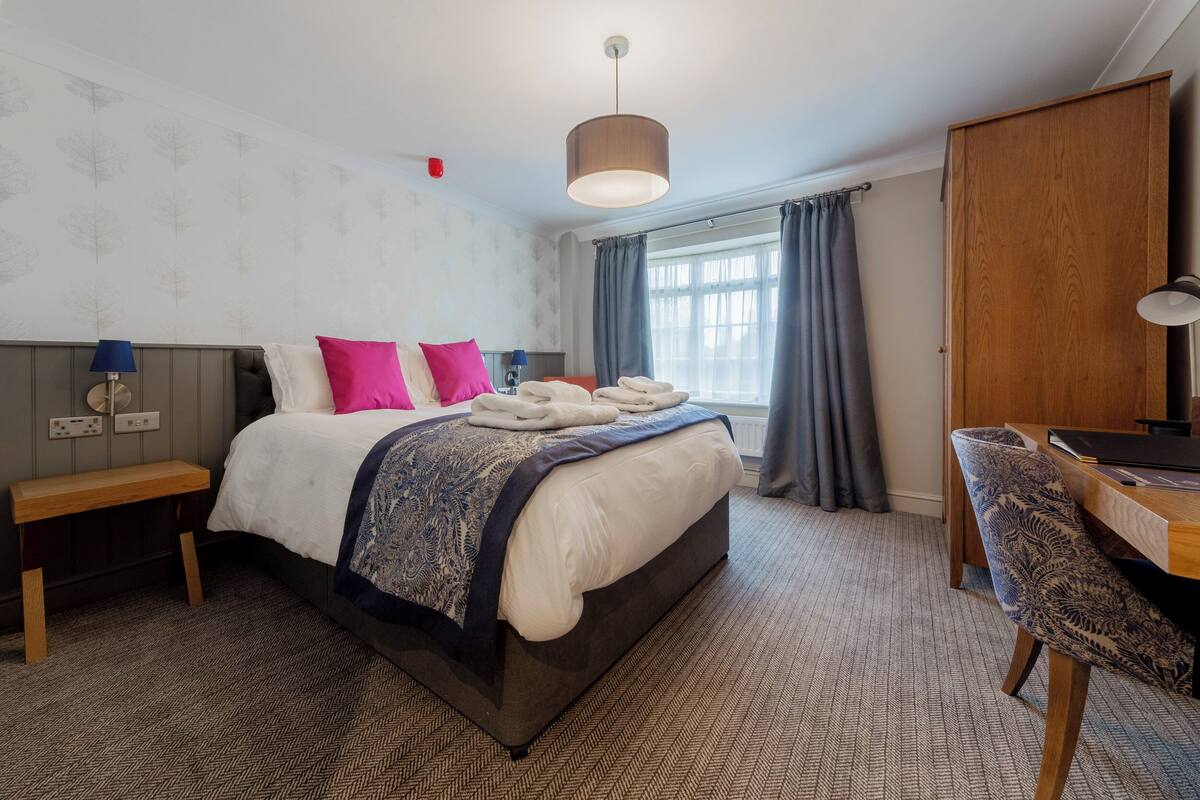 A hotel room with a large bed, a desk, a chair, a lamp, and a window with curtains.
