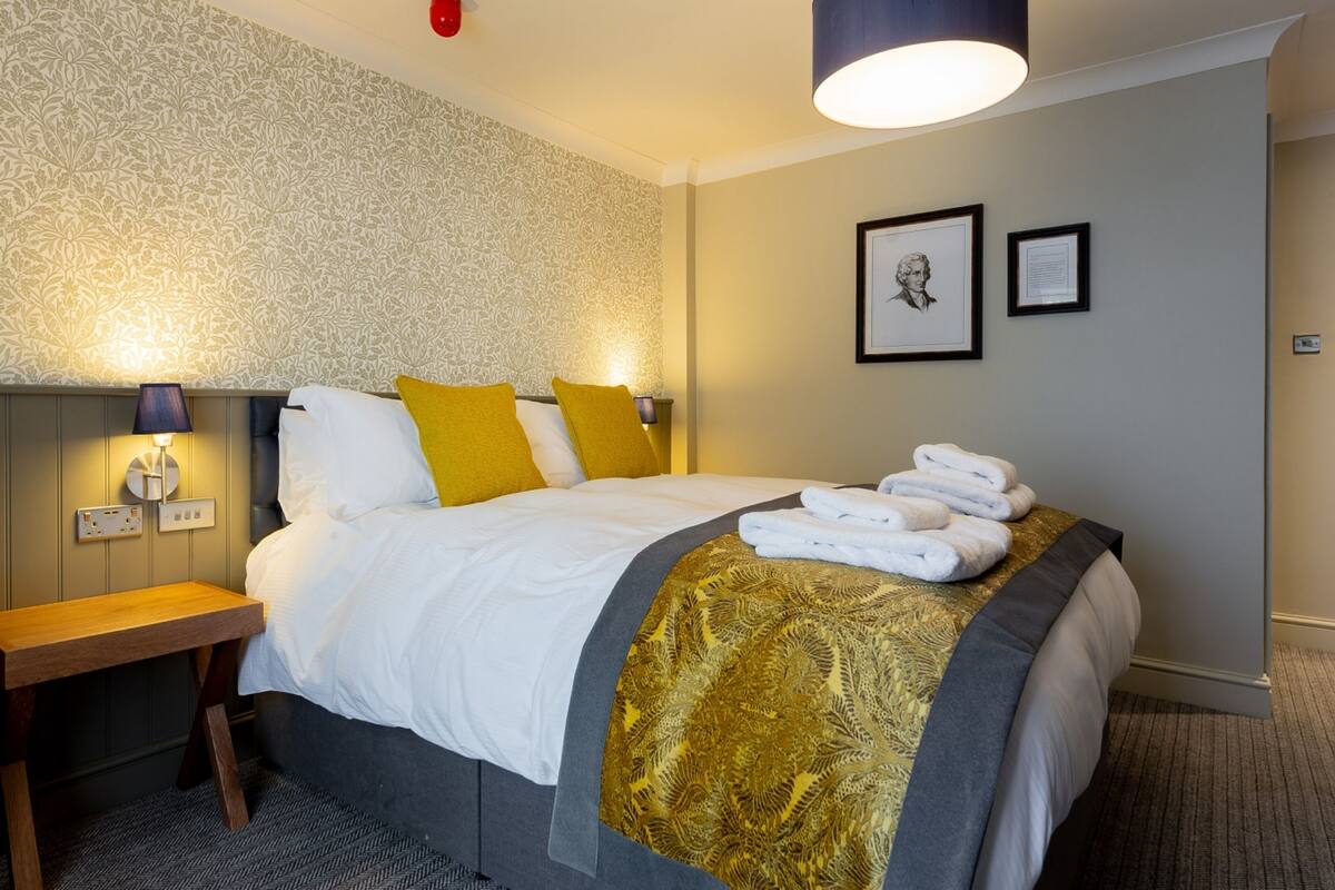 A neatly made bed with white linens and a gold-patterned throw, a wooden nightstand with a lamp, and a wall with framed artwork.