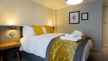 A neatly made bed with white linens and a gold-patterned throw, a wooden nightstand with a lamp, and a wall with framed artwork.