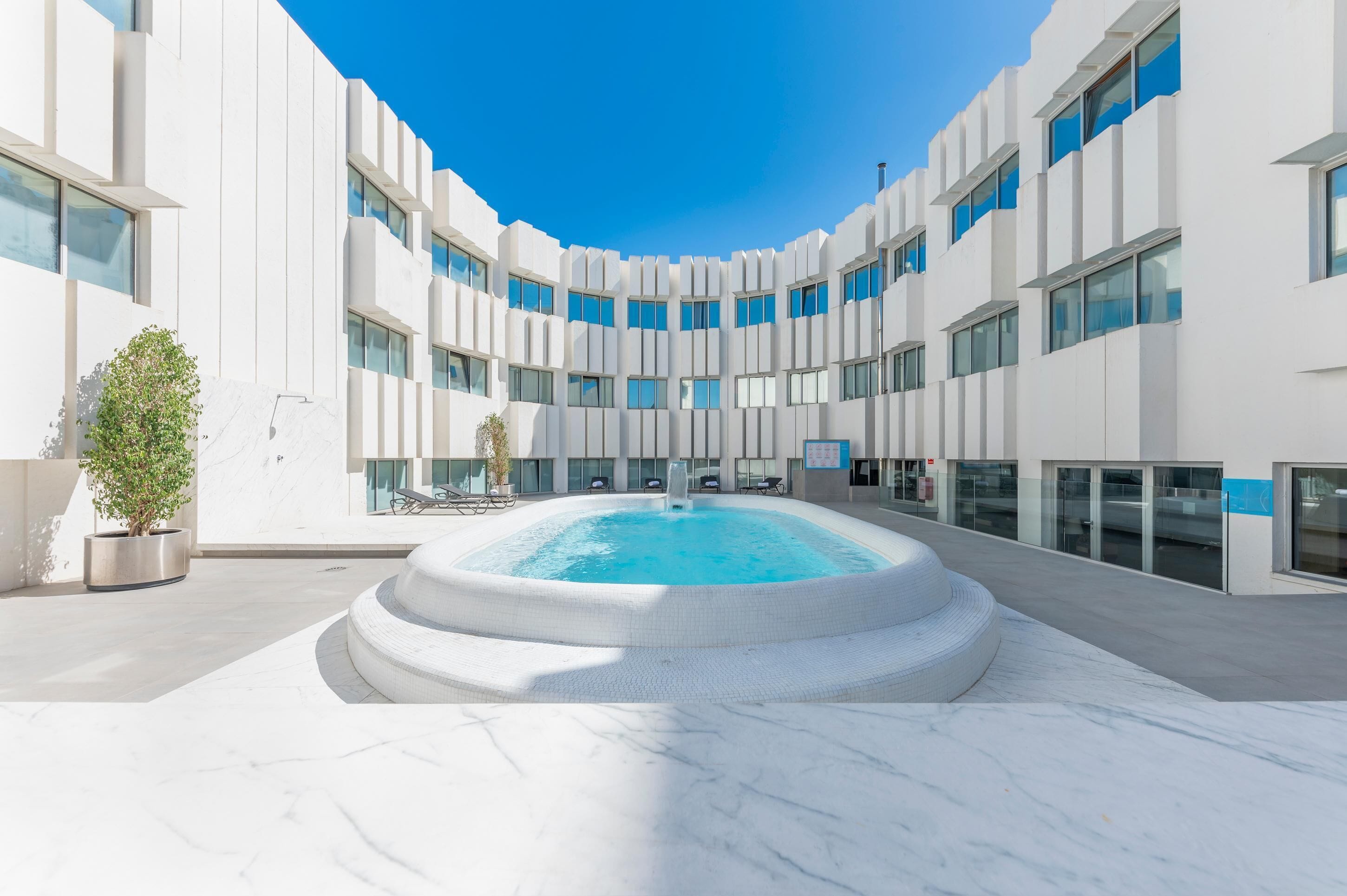 Un edificio moderno con piscina central, varios pisos y un cielo azul despejado.