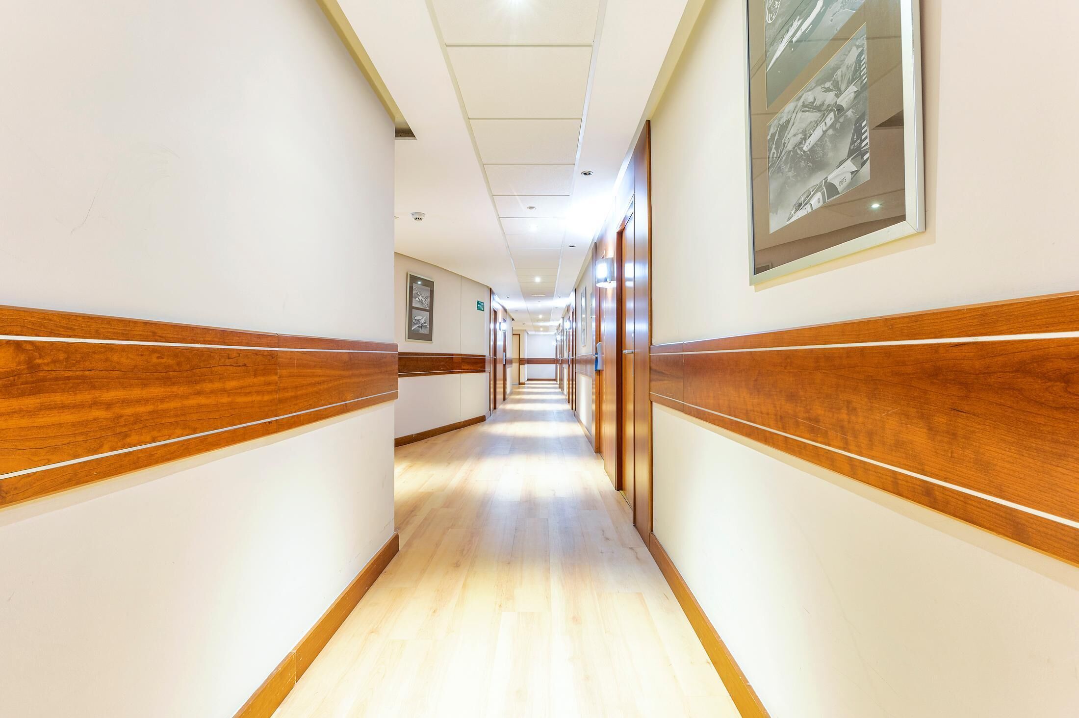 A hotel corridor with wooden paneling, framed pictures on the walls, and a light-colored floor.