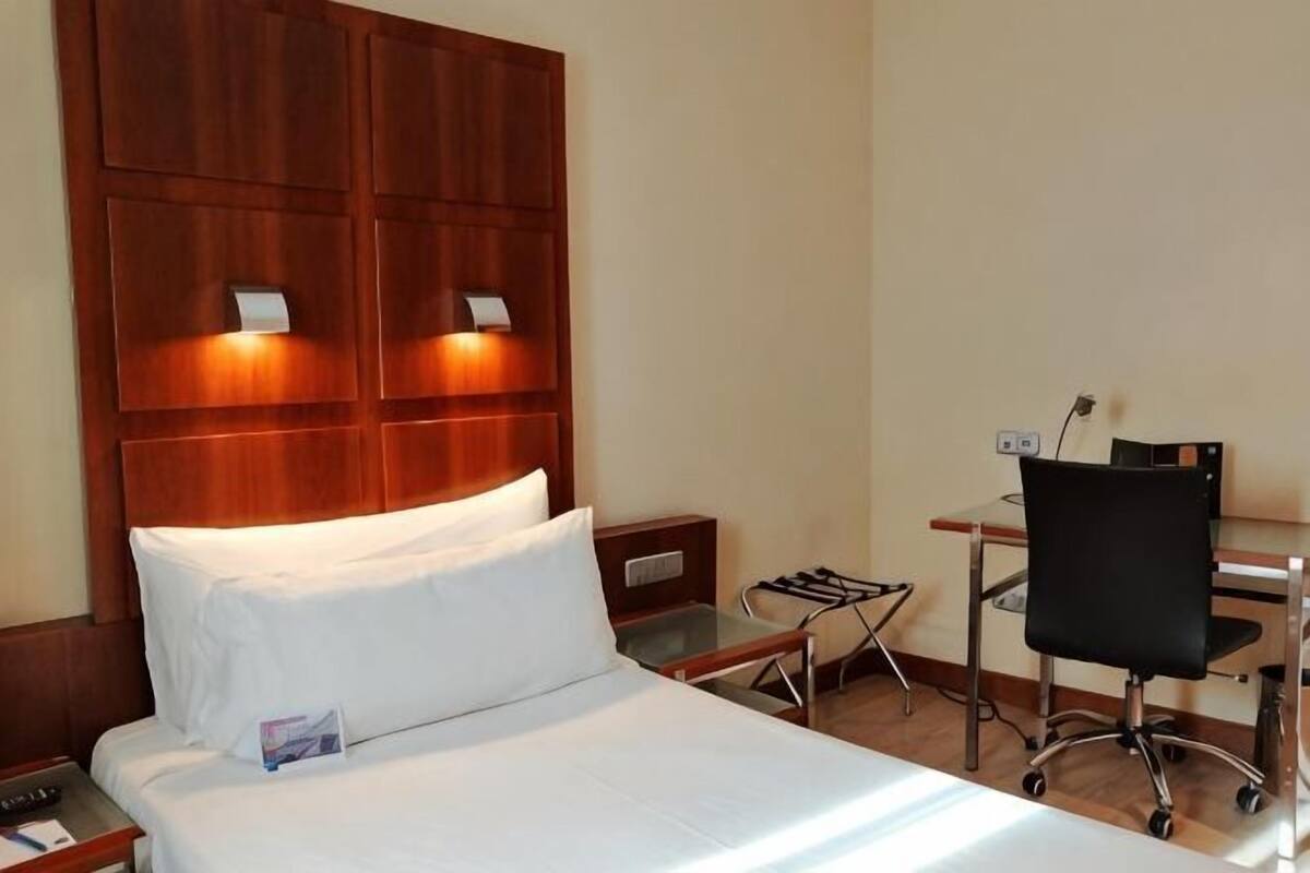 Hotelkamer met een houten hoofdeinde, een bed met witte lakens, een bureau met stoel en een nachtkastje.