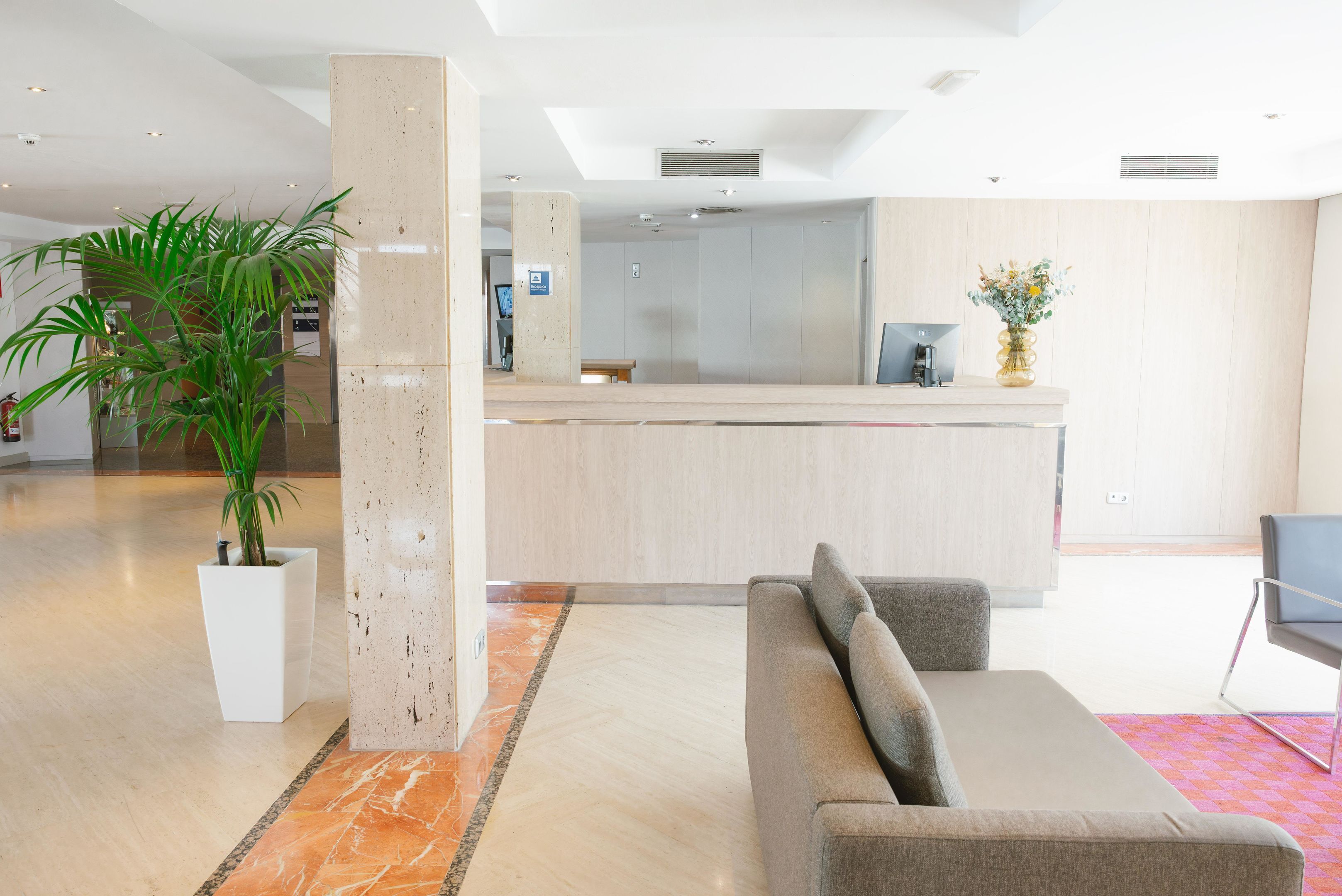 Een moderne lobby met een receptiebalie, een grijze bank, een grote potplant en een marmeren vloer.