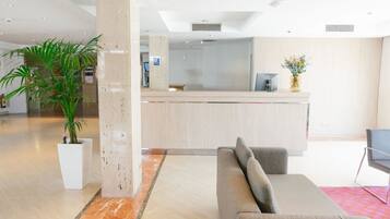 Een moderne lobby met een receptiebalie, een grijze bank, een grote potplant en een marmeren vloer.