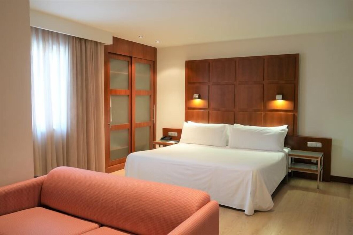 A hotel room with a large bed, a wooden headboard, two bedside tables with lamps, a sofa, and a sliding glass door.