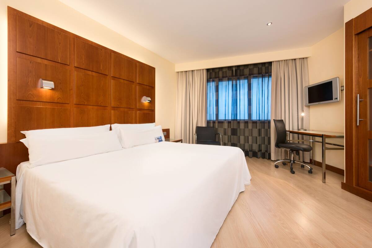Een hotelkamer met een groot bed, een bureau met stoel, een televisie en een raam met gordijnen.