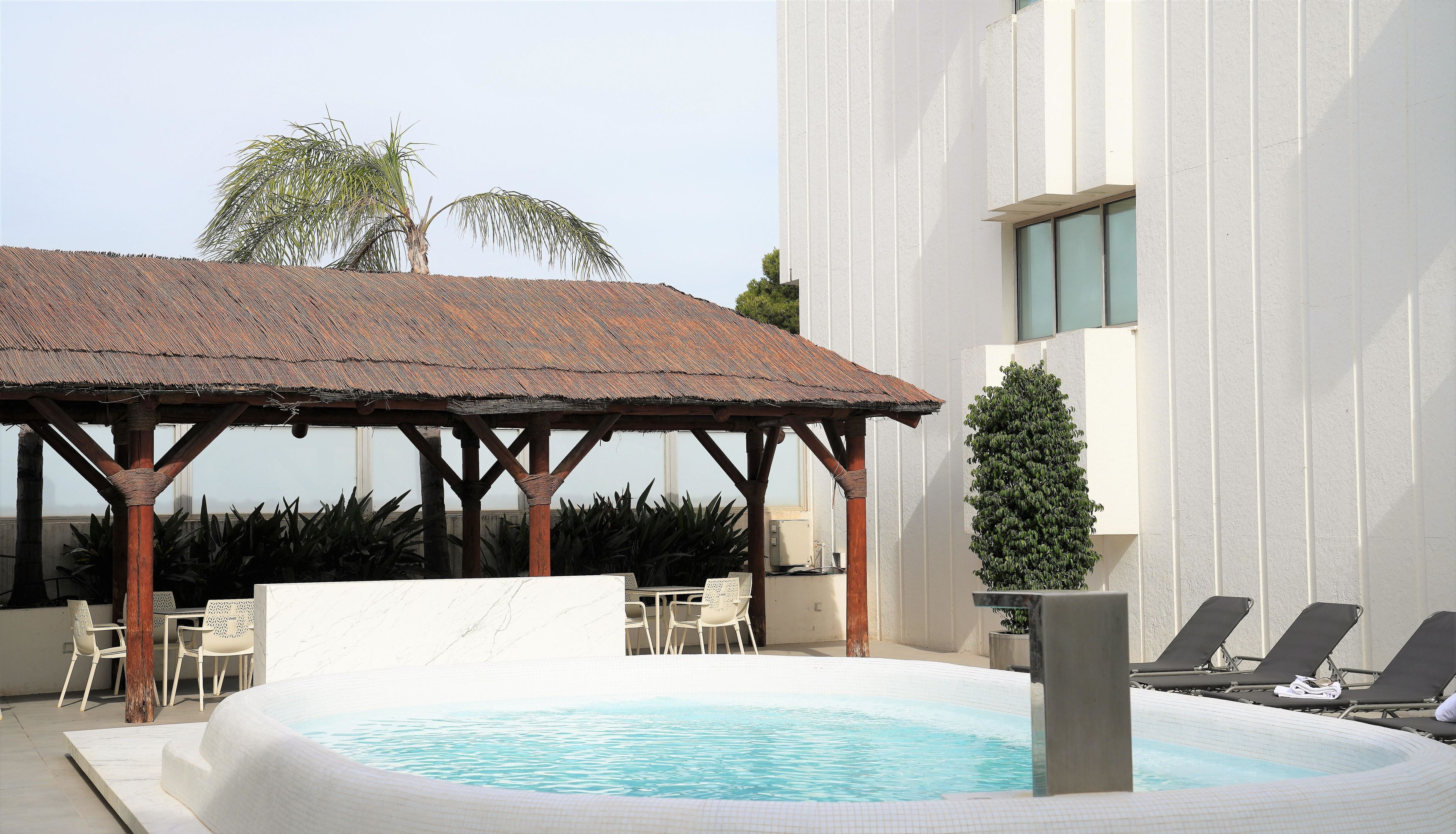 A pool area with a covered seating area, lounge chairs, and a small tree.