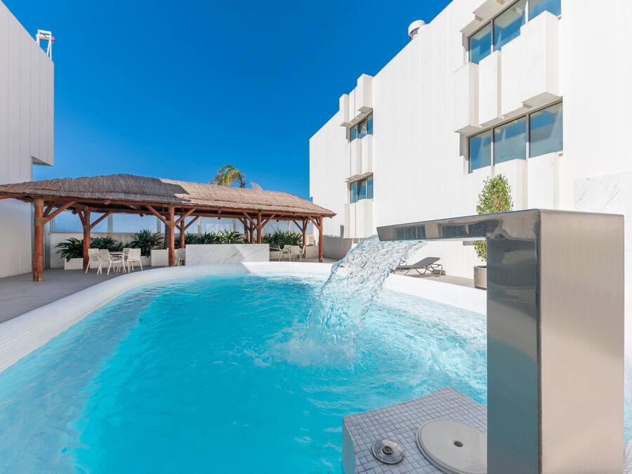 Una piscina con caída de agua, rodeada de edificios blancos y una estructura de techo de paja.