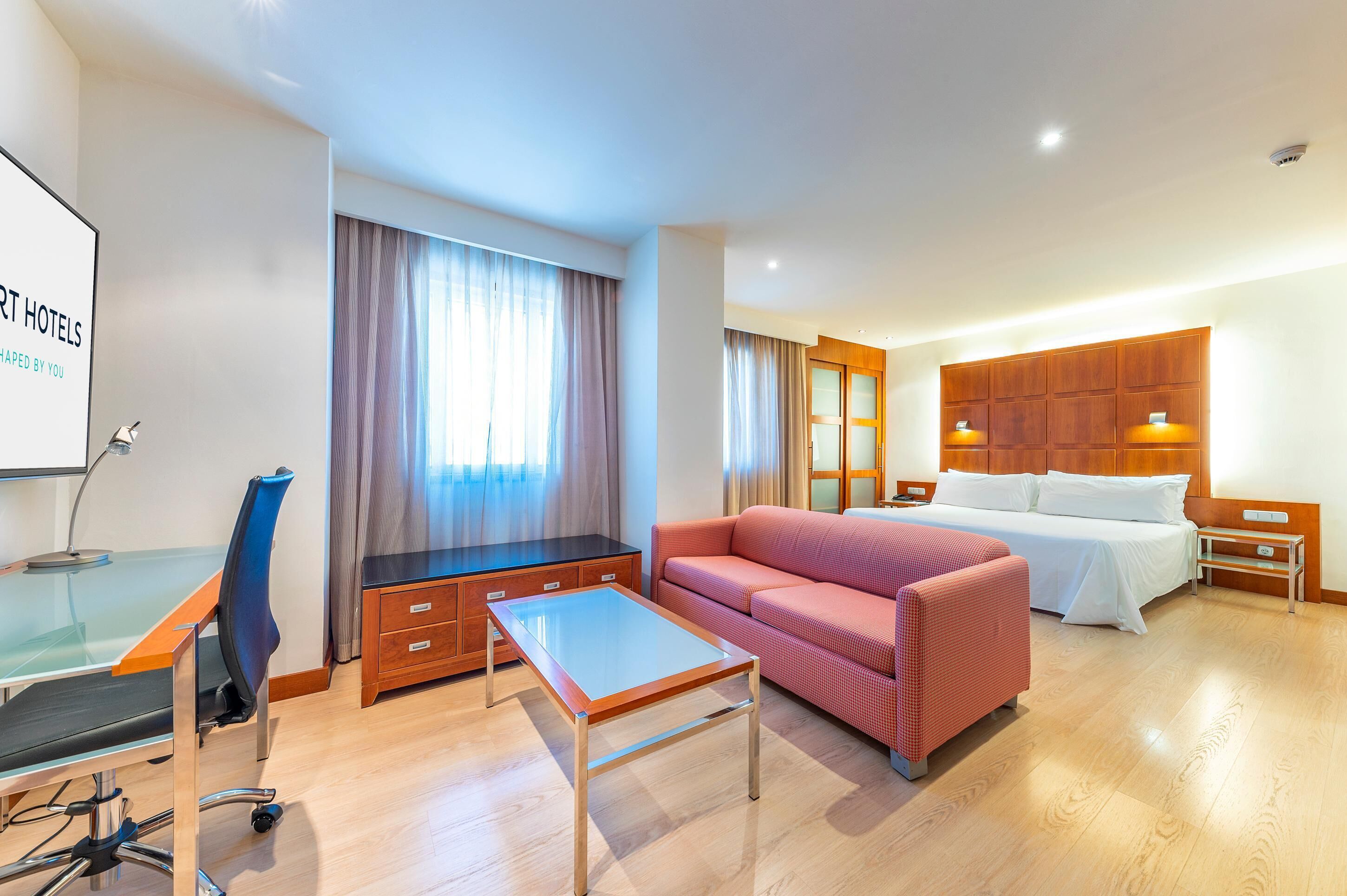 Una habitación de hotel moderna con un escritorio de vidrio, un sofá, una cama y un armario de madera.