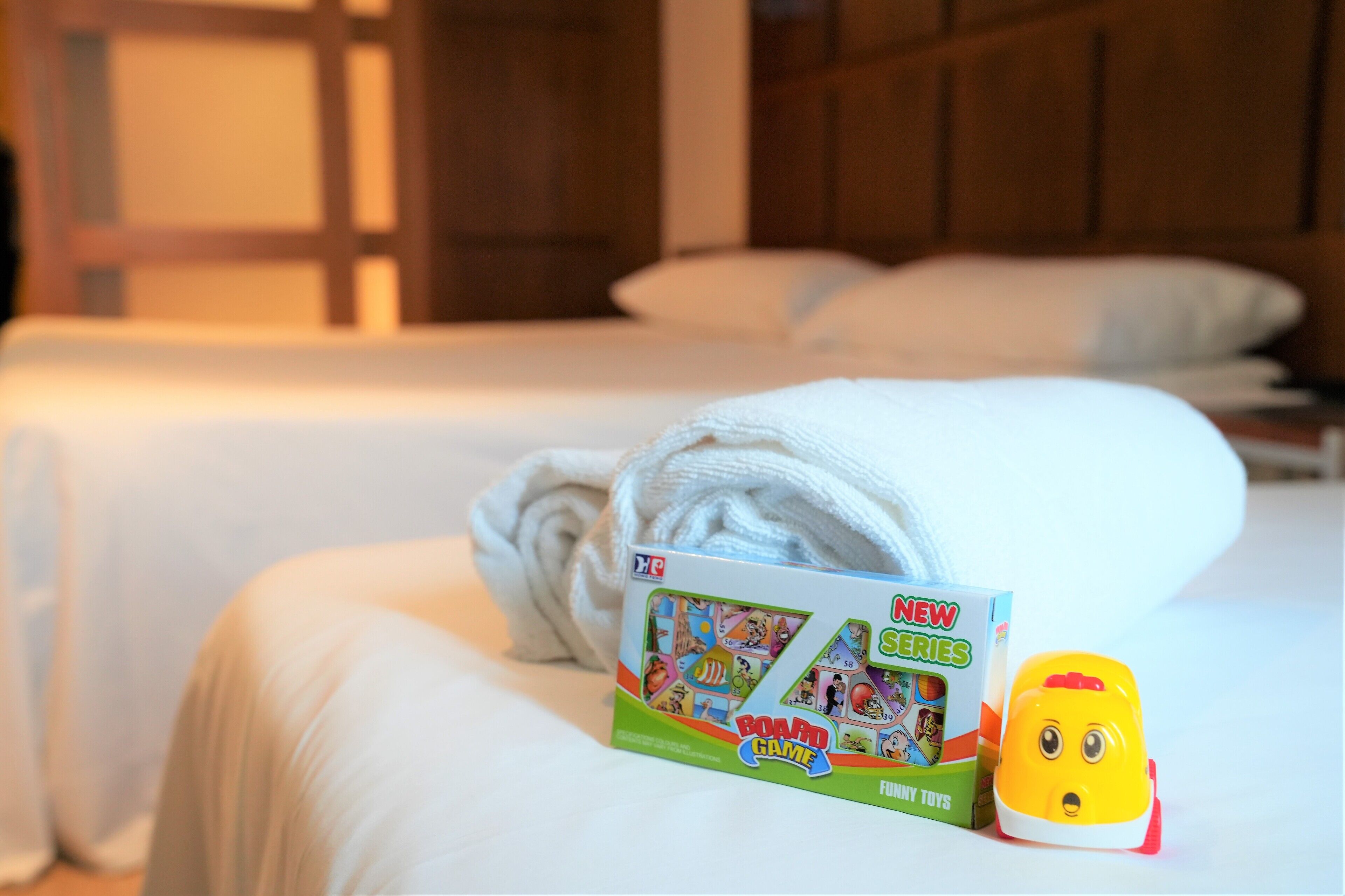 Una cama bien hecha con una toalla blanca enrollada, una caja de juego de mesa etiquetada "NEW SERIES" y un juguete pequeño sobre la cama.