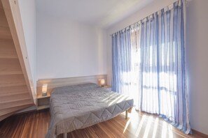 Un dormitorio con cama, suelo de madera, mesita de noche con lámpara y ventana con cortinas.