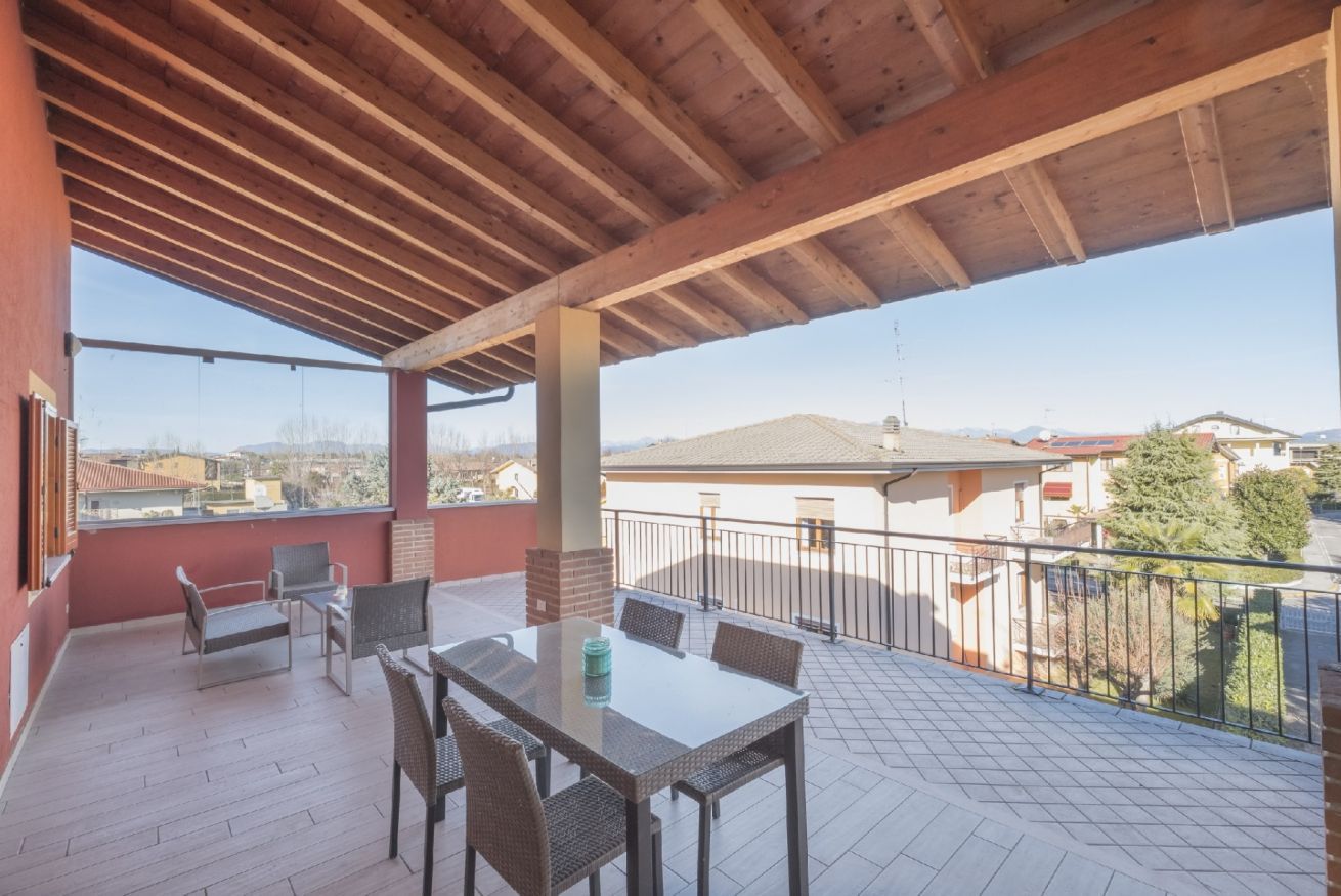 Une terrasse extérieure couverte, avec une table et des chaises, une cheminée en briques et une vue sur les bâtiments environnants.