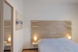 Un dormitorio con cama, persianas de madera, una lámpara de noche, un cuadro en la pared y un espejo.