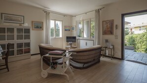 A living room with a wicker sofa, a small table, and a television. There are windows with curtains, a cabinet with glass doors, and a view of the outside.
