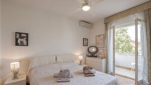A bedroom with a bed, bedside tables, a ceiling fan, an air conditioning unit, a window with curtains, and a view of the outside.