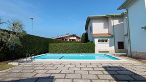 Una piscina con un borde pavimentado, setos y un edificio con techo rojo al fondo.