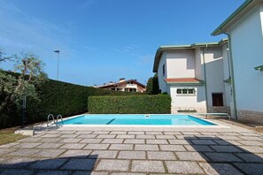 Una piscina con un borde pavimentado, setos y un edificio con techo rojo al fondo.