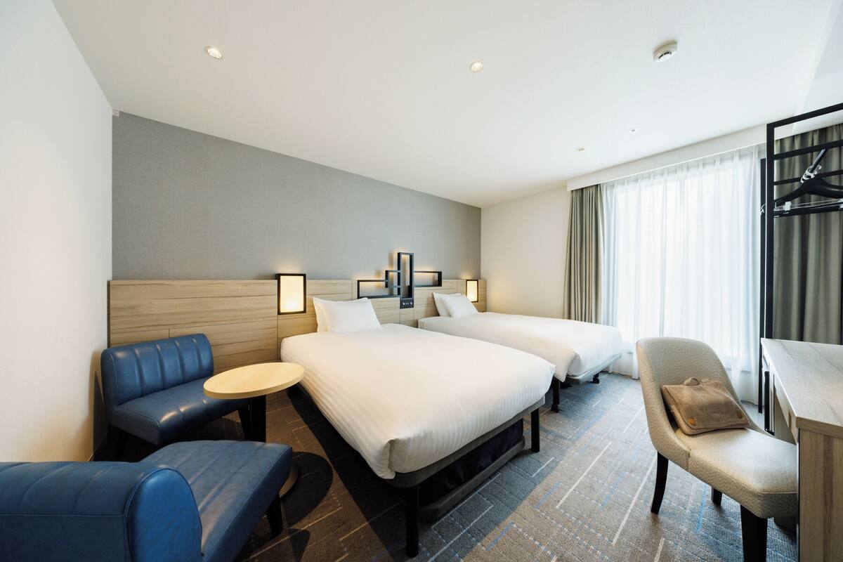Ein Hotelzimmer mit zwei Betten, einer Sitzecke, einem Schreibtisch und einem Fenster mit Vorhängen.