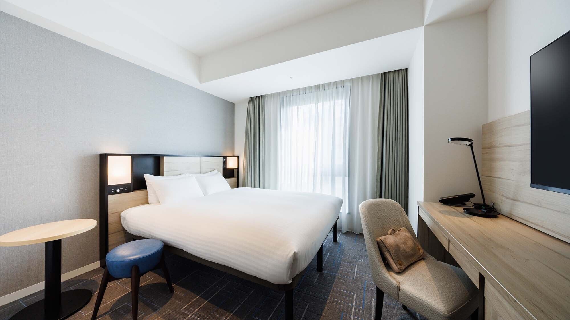 A hotel room with a large bed, a desk, a chair, a lamp, and a TV.