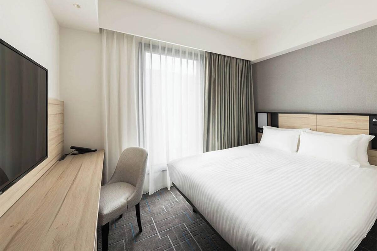 Ein Hotelzimmer mit Bett, Schreibtisch, Stuhl, Fernseher und einem Fenster mit Vorhängen.