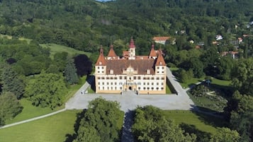 Standing proudly on a hill, this historic building boasts a grand facade with multiple towers and a central entrance, surrounded by lush greenery.