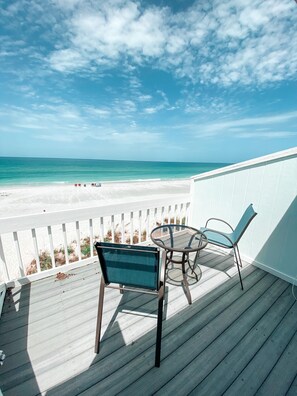A wooden deck with two chairs and a table, overlooking a beach with a white fence and a clear sky. - On the beach,Pool,Pet Friendly,Tennis/Pickleball,Kayaks,Paddle boards,Bikes (Englewood)
