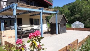 A covered patio area with a table and chairs, a small shed, and a garden with pink flowers. - Family chalet with sauna for 10-12 people (Gérardmer)