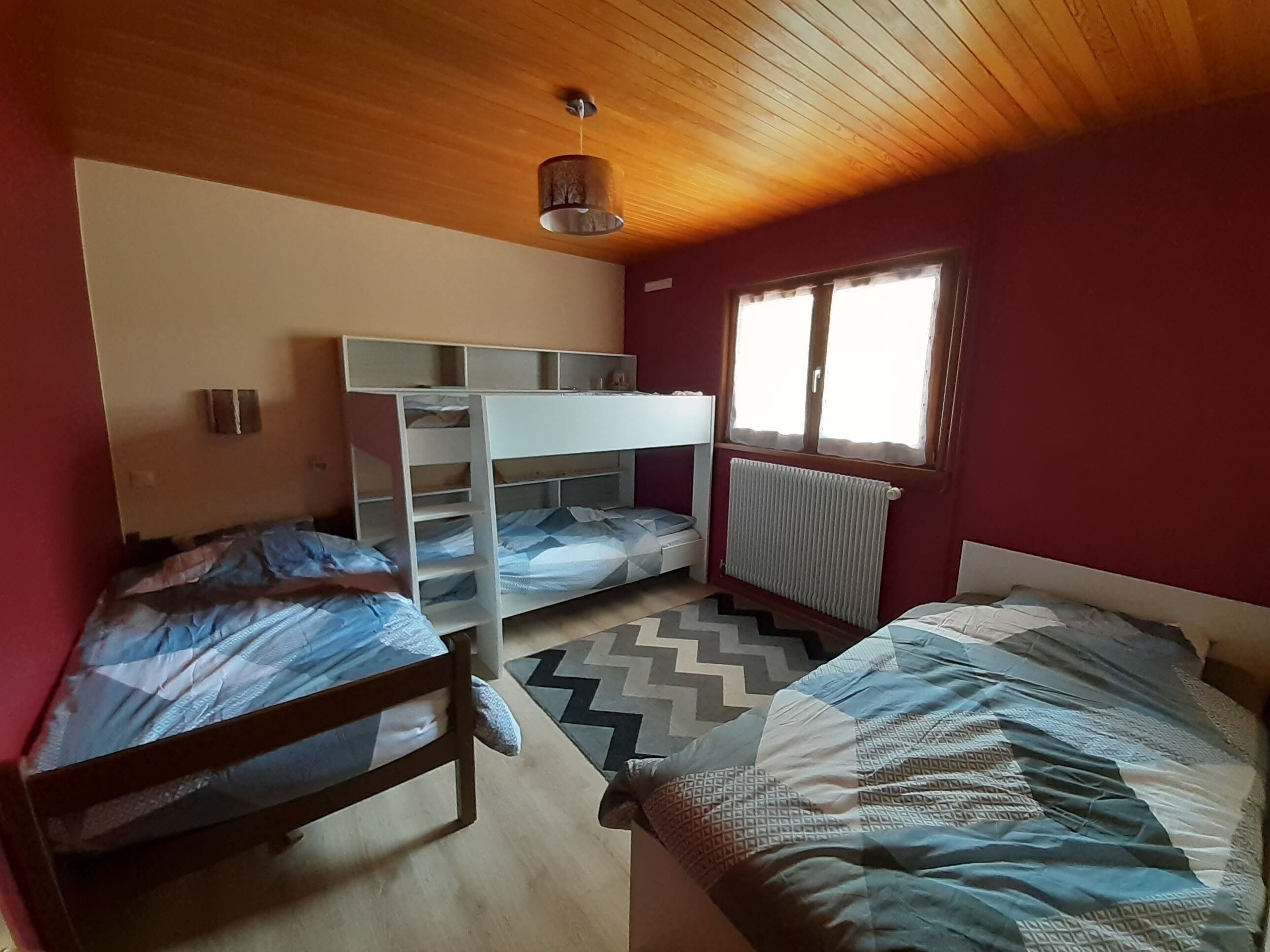 A dormitory room with two bunk beds, a wooden ceiling, a window with curtains, and a radiator.