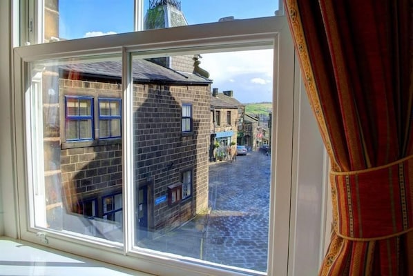 A window view of a cobblestone street with traditional stone buildings, a clear sky, and a curtain partially drawn to the side.