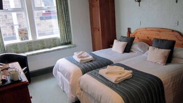 A double bed with two pillows and a folded towel on each side. There is a wooden headboard, a window with curtains, and a small table with a cup and saucer.