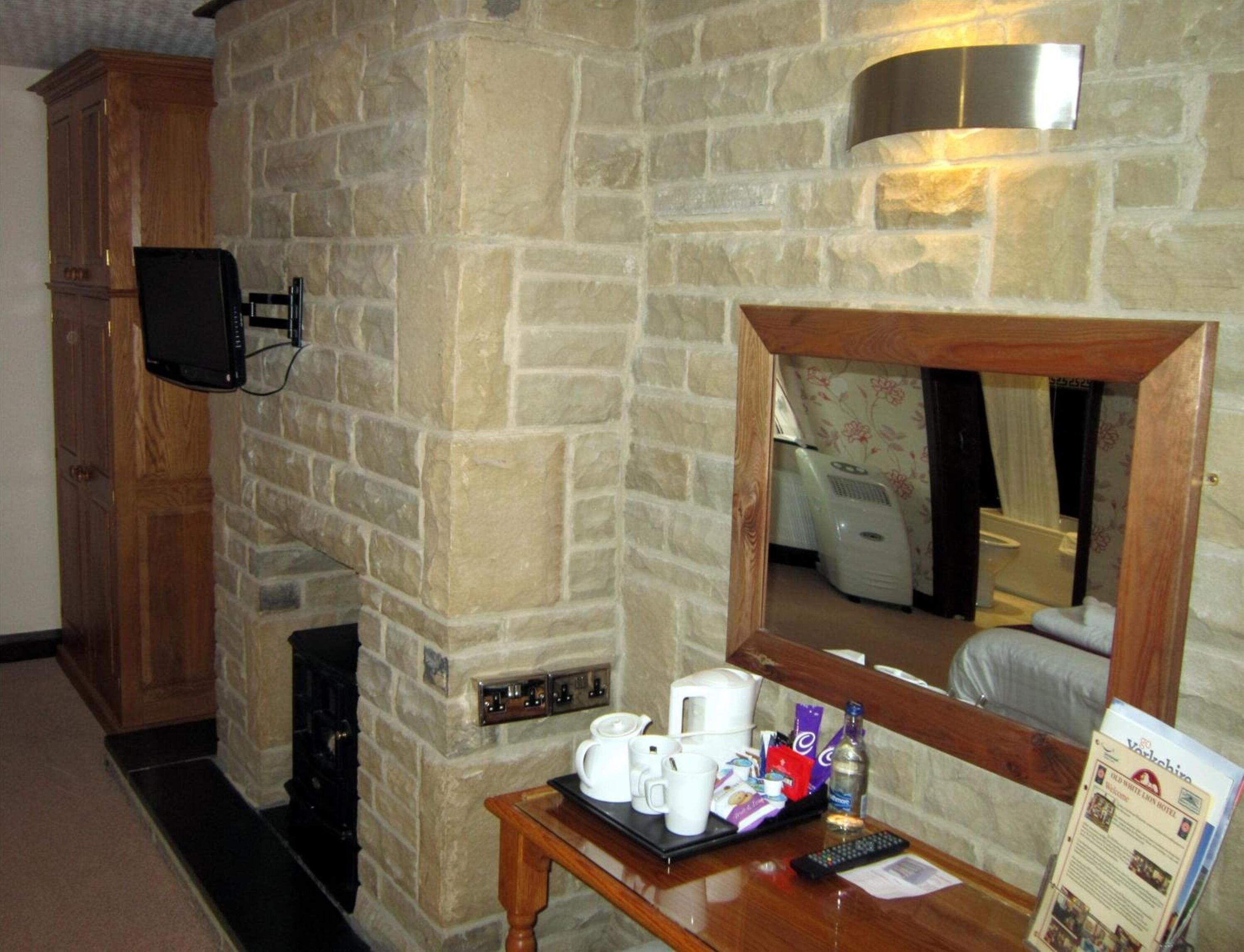 A stone-walled room with a wooden cabinet, a television mounted on the wall, a mirror, and a table with a tray of coffee cups and a remote control.