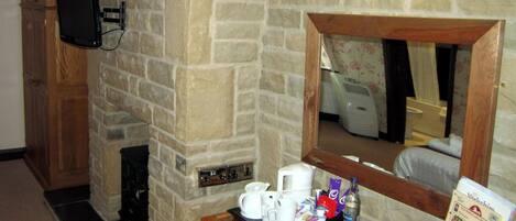 A stone-walled room with a wooden cabinet, a television mounted on the wall, a mirror, and a table with a tray of coffee cups and a remote control.