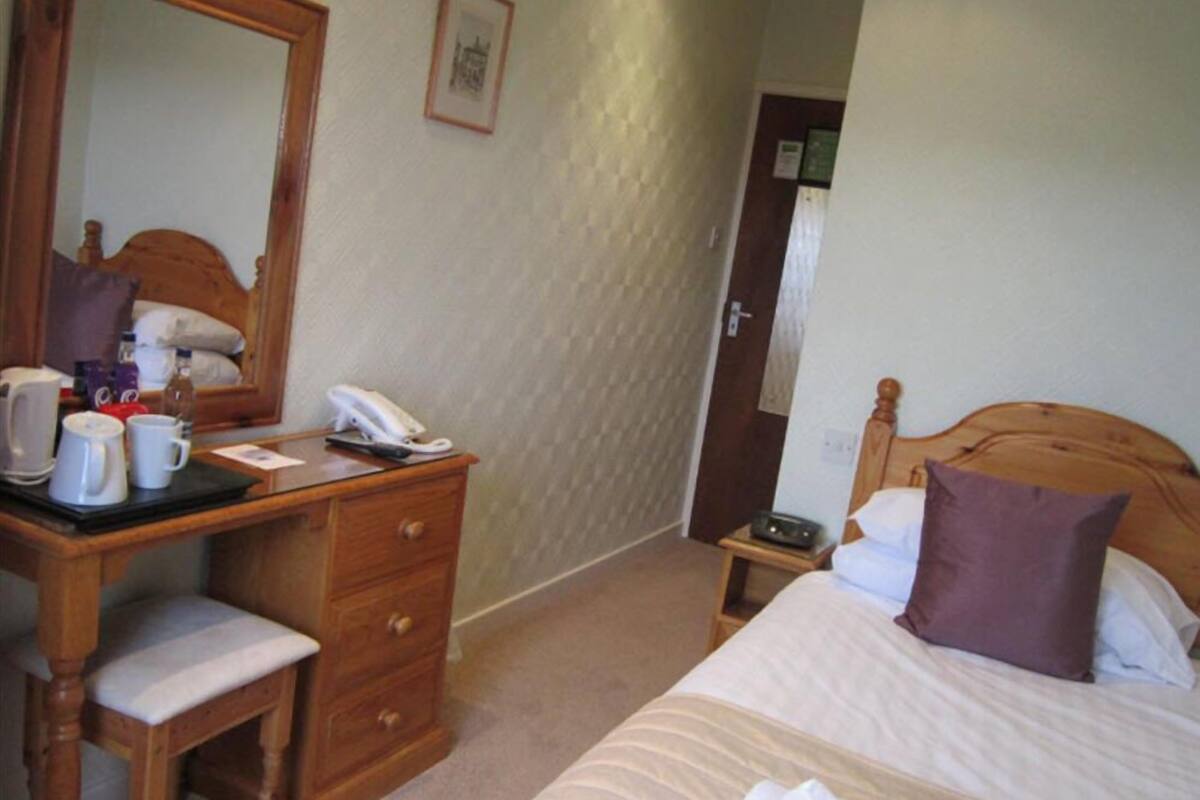 A hotel room with a wooden desk, a chair, a bed with a headboard, a bedside table, and a mirror.