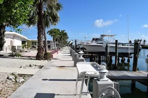 A marina with boats docked, a concrete walkway, palm trees, and a building with outdoor seating.