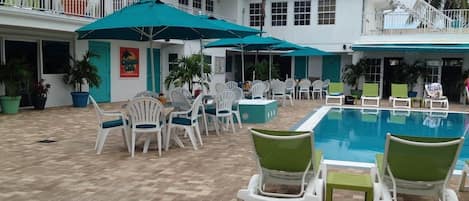 A pool area with white lounge chairs and green cushions, surrounded by a white building with multiple windows and a balcony.