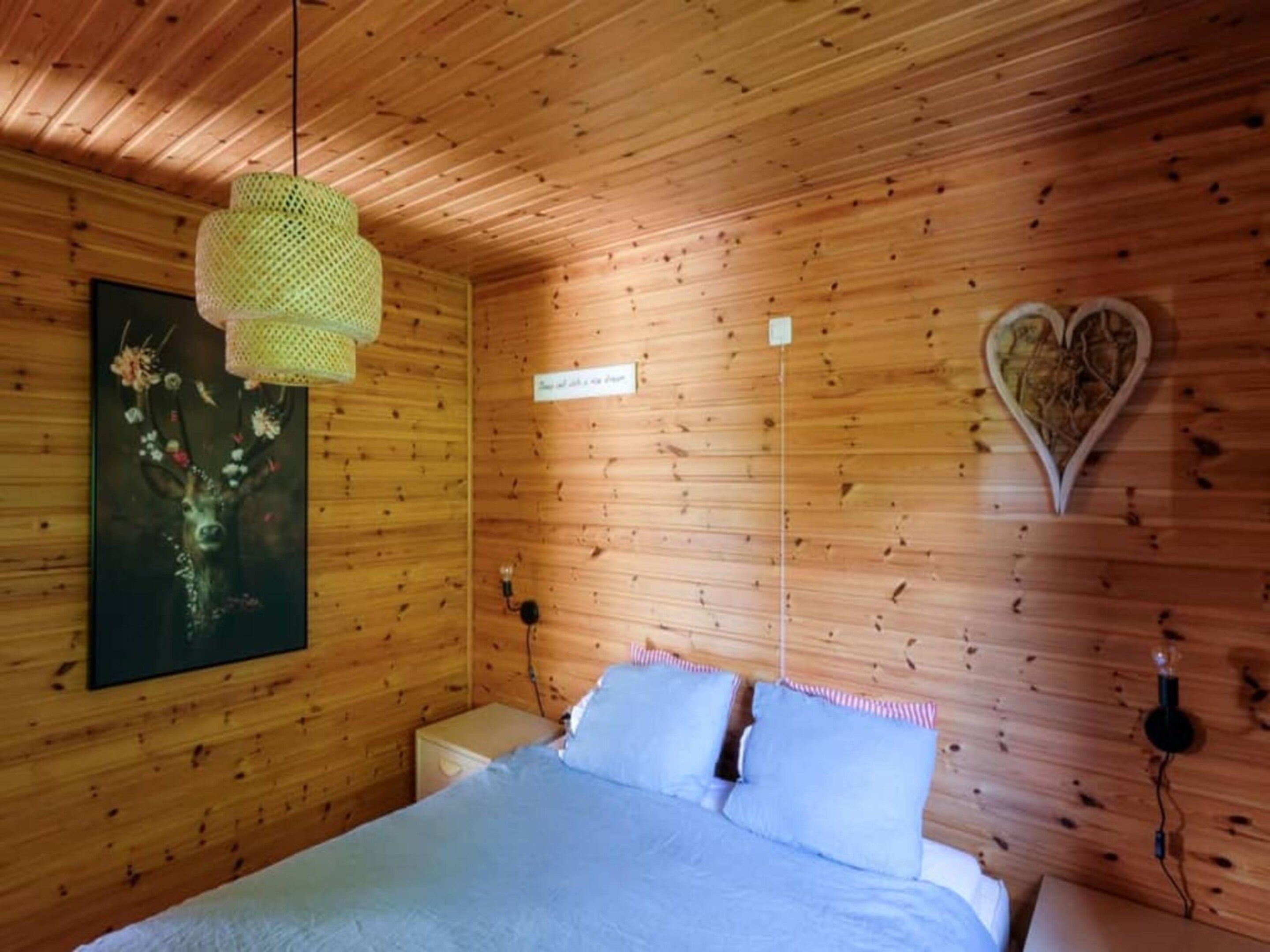 A wooden cabin room with a bed, a hanging light fixture, a wall-mounted heart-shaped decoration, and a framed picture.