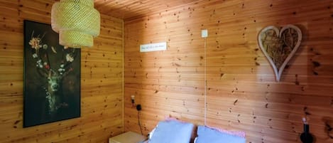 A wooden cabin room with a bed, a hanging light fixture, a wall-mounted heart-shaped decoration, and a framed picture.